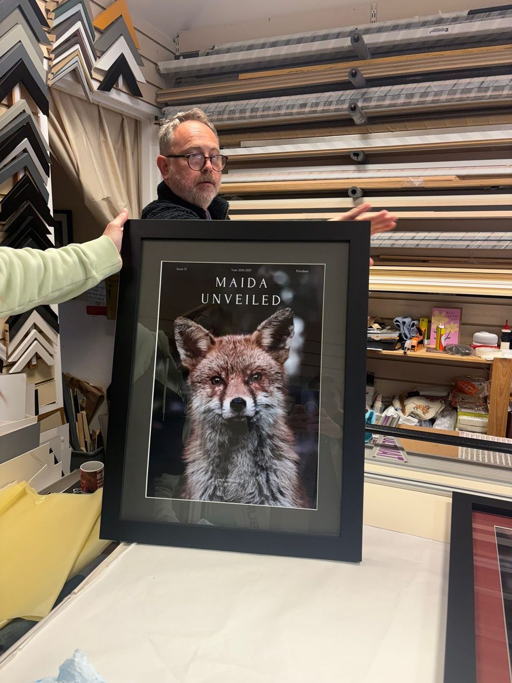 The Scottish master framer in his workshop, holding the finished framed print of Maida Fox among rows of frame mouldings